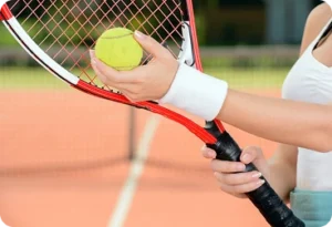 A close-up of a woman's hands holding a yellow tennis ball against the red strings of a tennis racket on a clay court.