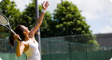 A young woman in a white tank top serving a tennis ball on an outdoor court with green trees in the background.